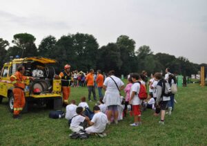 Due uomini in tuta arancione Protezione Civile vicini ad auto gialla insieme a bambini