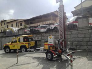 Auto gialla Protezione Civile e torre faro vicino a muretto di un parcheggio