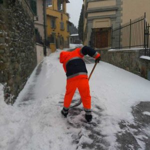 Persona della Protezione Civile in tuta arancione che spala neve su strada in salita tra le case